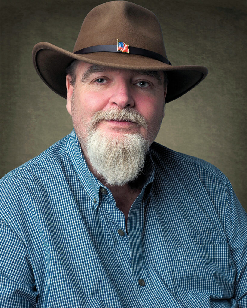 Professional portrait of M. Kameron Hawkins wearing a brown cowboy hat with an American flag pin and a blue checkered shirt, representing the M. Kameron Hawkins Foundation, Inc.